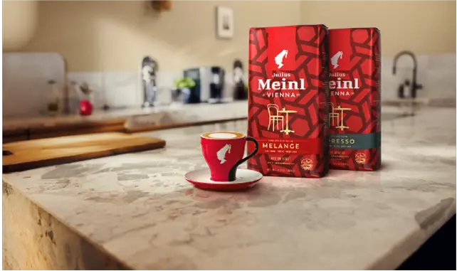Divas Julius Meinl kafijas paciņas un sarkana kafijas tasīte uz virtuves virsmas, fonā redzama mājīga virtuve.