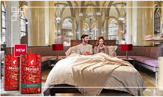 Pāris brokastīs gultā ar Julius Meinl kafiju elegantā interjerā; priekšplānā redzamas divas Julius Meinl kafijas iepakojuma paciņas.