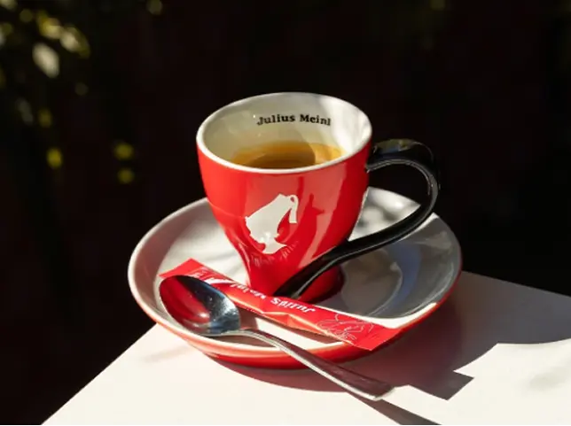 Sarkana Julius Meinl kafijas tasīte ar apakštasīti un karoti uz balta galda