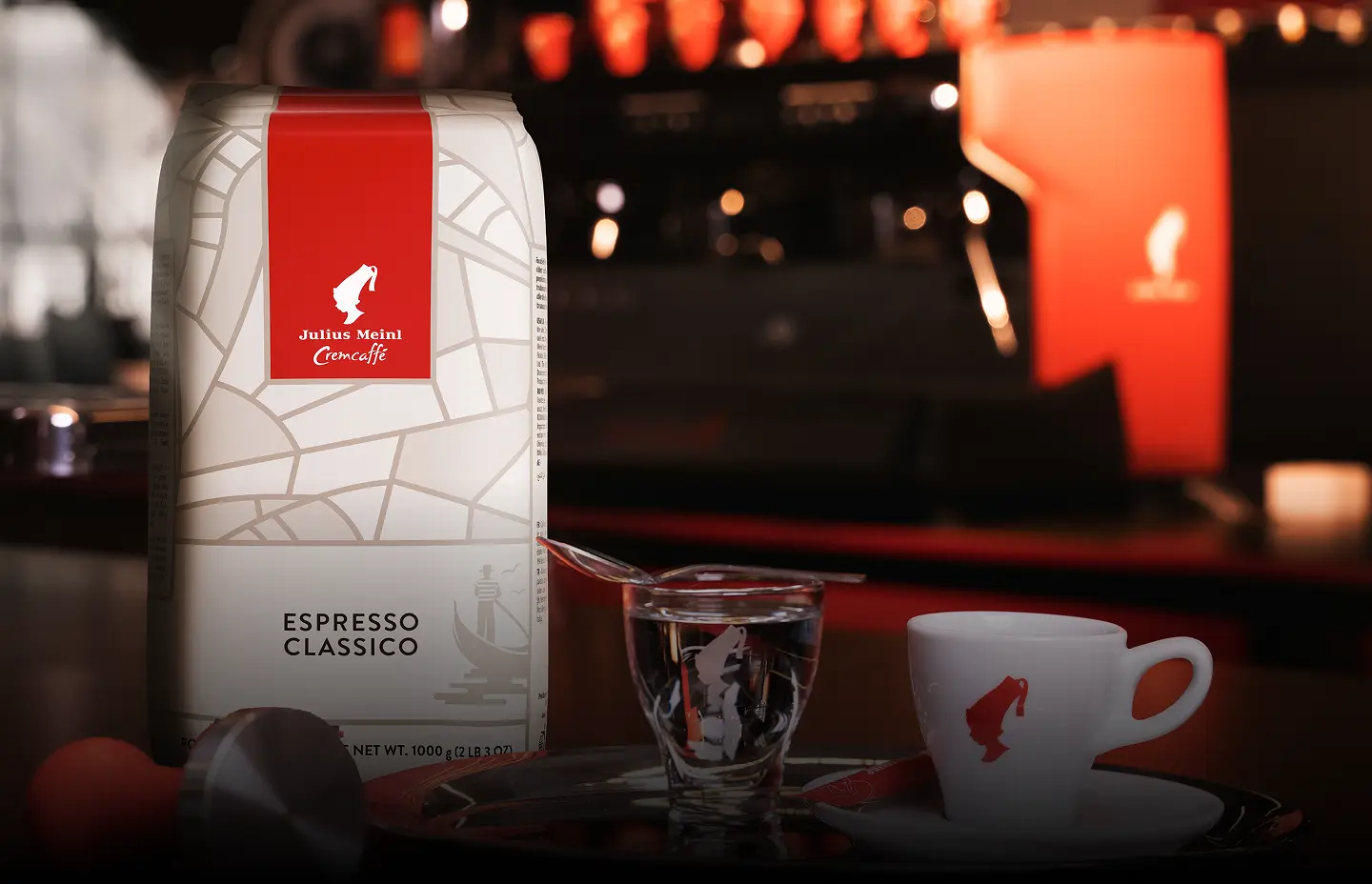 Julius Meinl “Espresso Classico” kafijas iepakojums uz koka letes, blakus mazā tasīte, ūdens glāzīte un metāla karotīte, fonā daļēji redzams kafijas automāts un zīmola sarkanā krāsa.