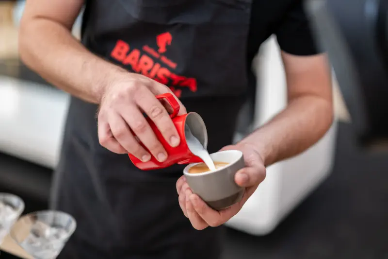 Pogas fona attēls “Kafejnīcām”: barista lej pienu espresso tasītē kafejnīcas vidē