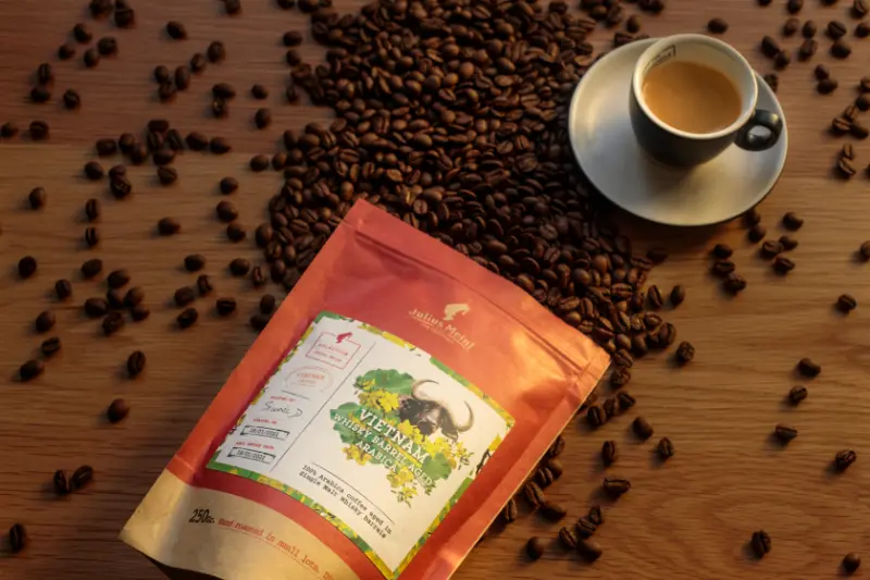 Pogas fona attēls “Veikaliem”: kafijas graudi izkaisīti ap Julius Meinl iepakojumu un tasīti uz koka virsmas
