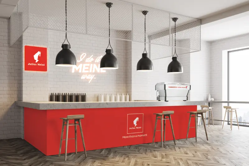 Pogas fona attēls “Viesnīcām”: Julius Meinl kafijas letes zona viesnīcas foajē ar sarkanu joslu un logotipu